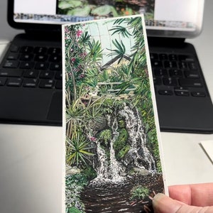 May include: A detailed illustration of a waterfall cascading into a pond, surrounded by lush greenery and tropical plants. The artwork features vibrant colours and intricate details, creating a serene and natural scene. The image is held in front of a blurred background.