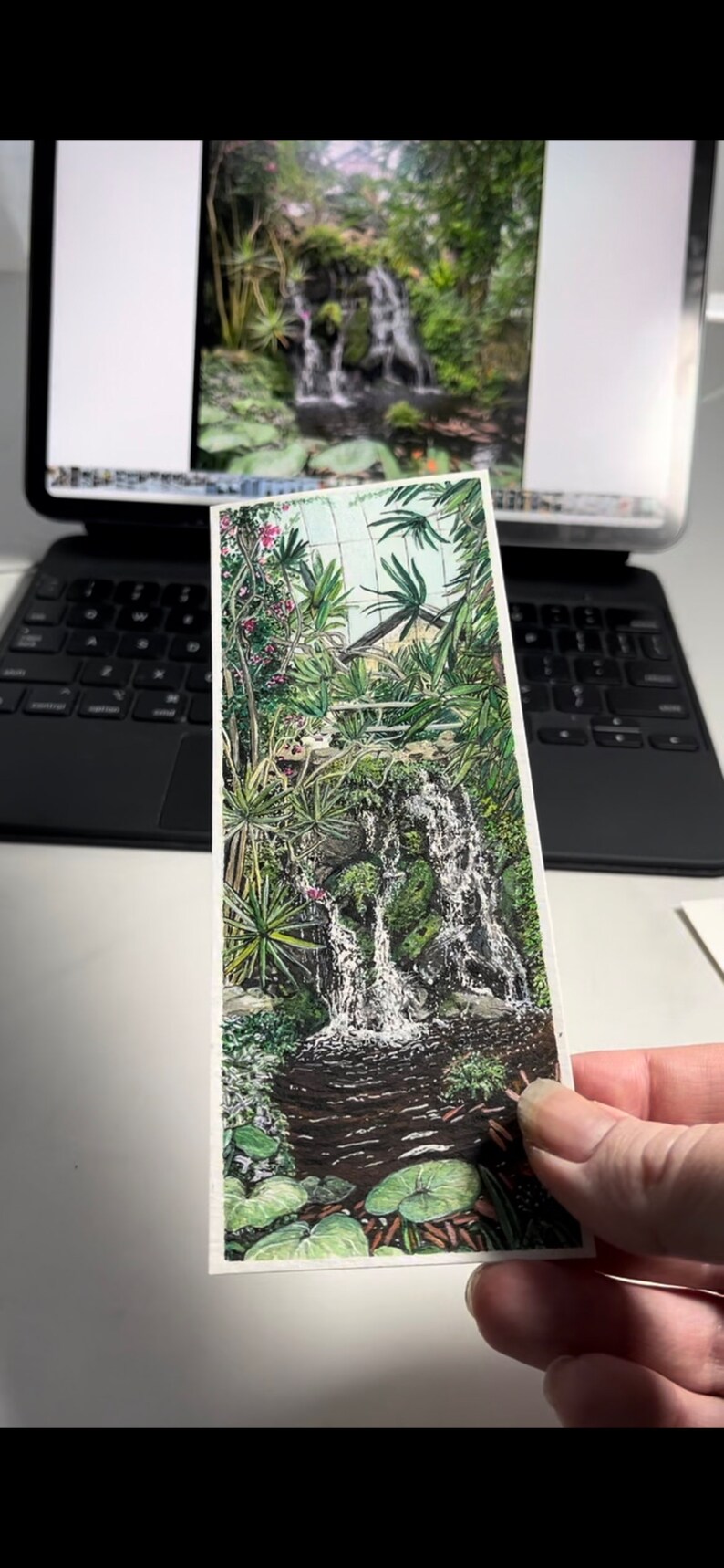 May include: A detailed illustration of a waterfall cascading into a pond, surrounded by lush greenery and tropical plants. The artwork features vibrant colours and intricate details, creating a serene and natural scene. The image is held in front of a blurred background.