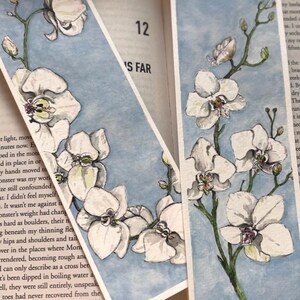 May include: Two watercolour bookmarks depicting white orchid flowers against a pale blue backdrop. The orchids have green stems and leaves. The bookmarks are on an open book.
