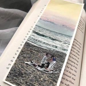 May include: A watercolour bookmark showing a beach scene with a couple. The sky fades from yellow to pink and blue. The couple is kneeling on the beach. The bookmark is inside an open book.