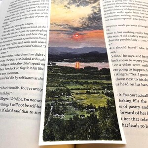 May include: A rectangular bookmark with a watercolour landscape painting. The artwork shows a sunset over a lake, with mountains and a forested area. The bookmark is between the pages of an open book.