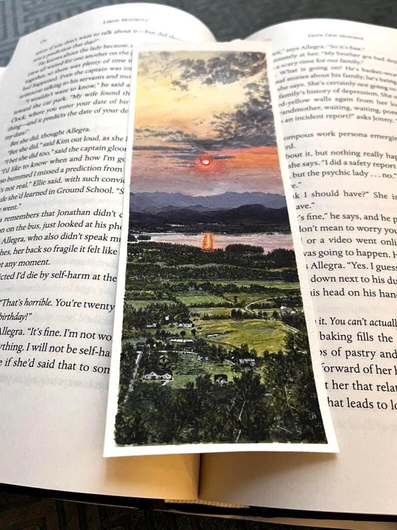 May include: A rectangular bookmark with a watercolour landscape painting. The artwork shows a sunset over a lake, with mountains and a forested area. The bookmark is between the pages of an open book.