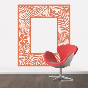 Pu&ograve; includere: Un adesivo da parete color corallo arancio con un design di foglie e fiori tropicali a forma di cornice. L'adesivo &egrave; su una parete bianca con una sedia rossa in primo piano.