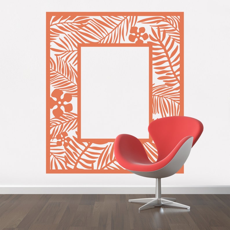 Pu&ograve; includere: Un adesivo da parete color corallo arancio con un design di foglie e fiori tropicali a forma di cornice. L'adesivo &egrave; su una parete bianca con una sedia rossa in primo piano.