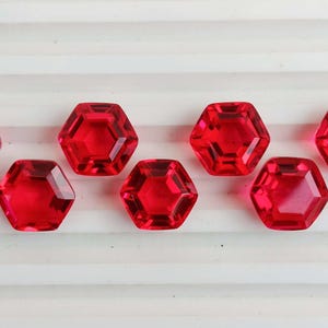 May include: Seven faceted, hexagonal, bright red gemstones are arranged in a row. The gems have a high-gloss finish and are set against a white, ridged surface. The gems are evenly spaced.