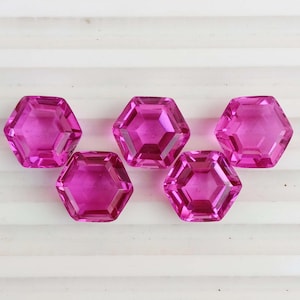 May include: Five faceted, hexagonal, hot pink gemstones are arranged on a white, ridged surface. The gems have a high degree of clarity and reflect light, showcasing their multiple facets. The gems are a vibrant shade of pink.