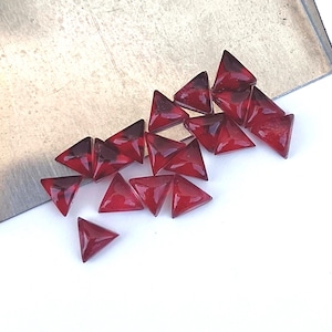 May include: A pile of small, red, triangular glass gems. The gems are scattered on a white surface.