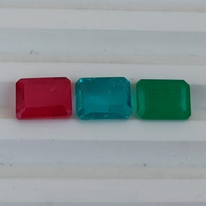 May include: Three rectangular gemstones in a row: red, teal, and green. Each gem has a faceted cut and a glossy finish. The gems are displayed on a white, ridged surface.