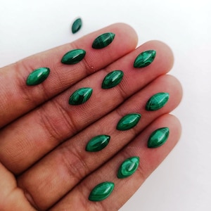 Puede incluir: Colección de gemas de malaquita con corte marquesa, cada una mostrando un color verde oscuro intenso con patrones arremolinados únicos. Las piedras pulidas se exhiben sobre un fondo claro, resaltando su belleza natural y diseños intrincados.