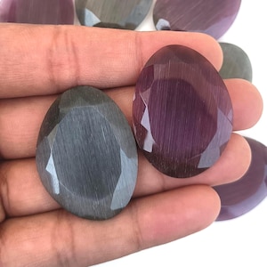 Puede incluir: Se muestran gemas ovaladas en tonos grises y morados. Las piedras tienen un corte facetado, que atrapa la luz y revela su estructura interna. Las gemas se sostienen en una mano, mostrando sus variaciones de tamaño y color.