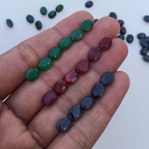 May include: A collection of oval-shaped gemstones in three colours: green, red, and blue. The gemstones are arranged in rows on a hand.