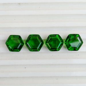 May include: Four faceted, hexagonal, emerald green gemstones are arranged in a row. The gems have a high degree of clarity and reflect light, showcasing their polished surfaces. The gemstones are likely for jewelry making.
