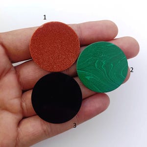 Peut inclure: Trois pierres circulaires dans une main. La pierre supérieure est d'un orange rougeâtre texturé. La pierre de droite est verte avec un motif tourbillonnant. La pierre inférieure est noire et brillante. Ces pierres pourraient être utilisées pour des bijoux ou des travaux manuels.