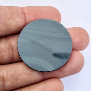 May include: A round, polished stone with a swirling pattern of gray and teal hues. The stone is held in a hand, showcasing its smooth surface and natural variations. The stone's circular shape is clearly defined.