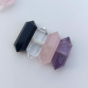May include: Four double-terminated crystal wands in a row. The crystals are black, clear, pink, and purple. The crystals are faceted and have a pointed end on each side. The crystals are arranged diagonally on a white surface.