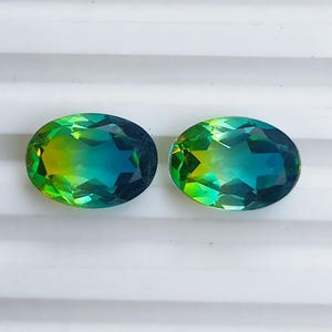 May include: Two oval-cut gemstones with a gradient of colors. The gems transition from a vibrant yellow-green to a deep teal blue. The stones are set against a white background.