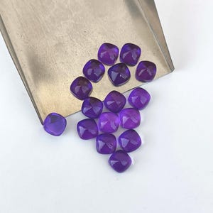 May include: A collection of faceted, square-cut, deep purple gemstones. The gems are arranged on a white surface with a silver-colored metal tray in the background. The gemstones have a polished, reflective surface.
