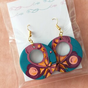 Puede incluir: Pendientes de mariposa de color turquesa y morado con ganchos dorados. El diseño de la mariposa está pintado en discos de madera redondos.