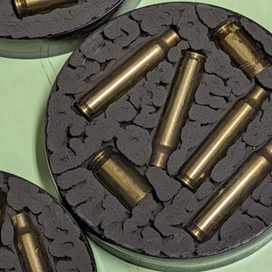 May include: Close-up of three round coasters. Each coaster features a dark gray, textured surface with brass bullet casings embedded within. The coasters are set against a light green background.
