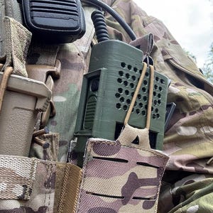 May include: An olive green tactical radio with a black microphone is attached to a camouflage vest. The radio has a series of ventilation holes and is secured in a camouflage pouch. A coiled cable connects the radio to the microphone.