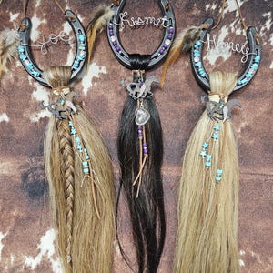 May include: Three decorative horseshoe ornaments with horse hair tassels. Each horseshoe is adorned with turquoise beads, feathers, and a wire name: "Joe", "Kismet", and "Honey". The background is a brown and white cowhide pattern.