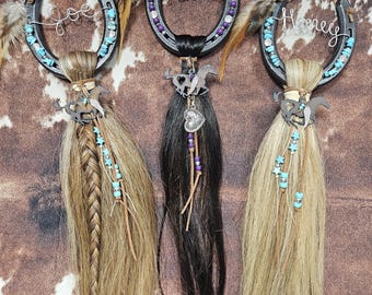 May include: Three decorative horseshoe ornaments with horse hair tassels. Each horseshoe is adorned with turquoise beads, feathers, and a wire name: "Joe", "Kismet", and "Honey". The background is a brown and white cowhide pattern.