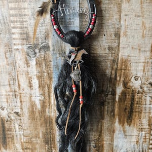 May include: A horseshoe-shaped wall hanging with the name "Harley" in wire. The horseshoe is decorated with red beads and a feather. Below the horseshoe is a bundle of black hair, a horse charm, and a heart charm.