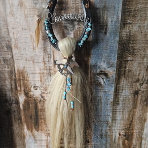 May include: A black horseshoe wall hanging with the word "Charm" in silver wire. Turquoise stones and a feather adorn the horseshoe. A bundle of blonde hair and a metal horse silhouette hang below, creating a rustic, Western-themed decoration.
