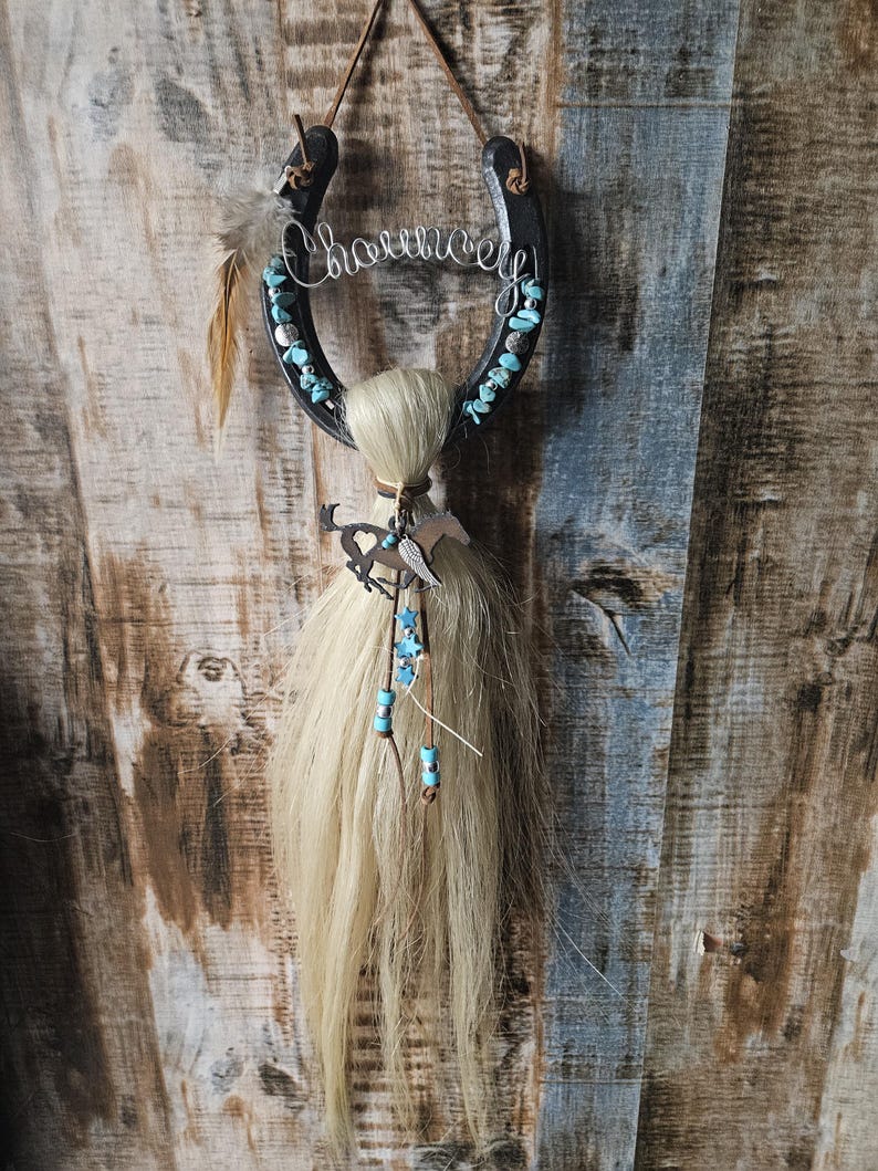May include: A black horseshoe wall hanging with the word "Charm" in silver wire. Turquoise stones and a feather adorn the horseshoe. A bundle of blonde hair and a metal horse silhouette hang below, creating a rustic, Western-themed decoration.