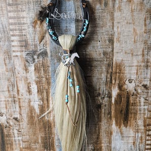 May include: A black horseshoe with the name "Sonna" in wire, adorned with turquoise stones and feathers. A blonde horsehair tassel hangs below, decorated with a silver horse charm and blue star beads. A rustic, country-themed wall hanging.