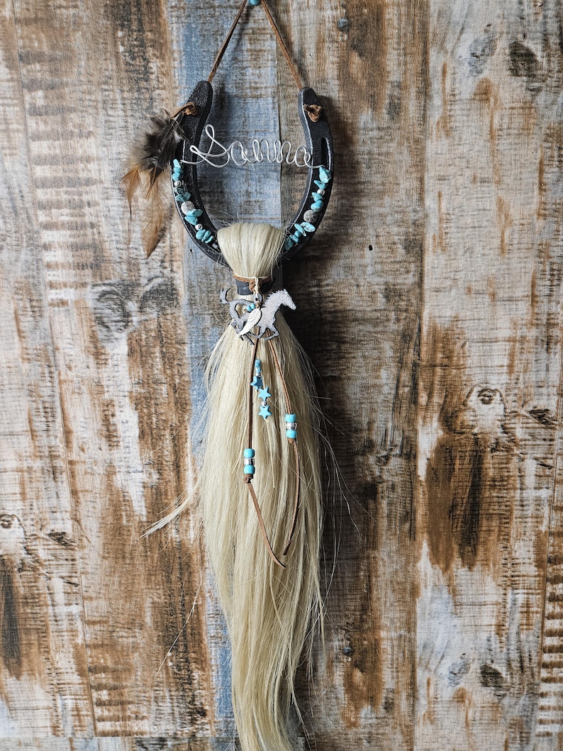 May include: A black horseshoe with the name "Sonna" in wire, adorned with turquoise stones and feathers. A blonde horsehair tassel hangs below, decorated with a silver horse charm and blue star beads. A rustic, country-themed wall hanging.