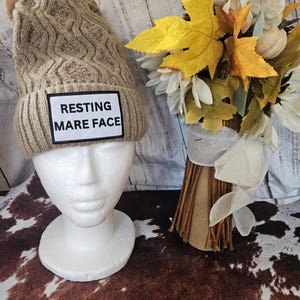 May include: A tan cable knit beanie with a faux fur pom-pom. The beanie has a rectangular patch with the words "RESTING MARE FACE" in black text. The beanie is displayed on a white mannequin head.