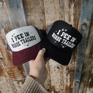 May include: Two trucker hats with a white mesh back and a black front. The hats have the text "I pee in horse trailers" in white with stars on either side of the text. One hat is burgundy and the other is black.