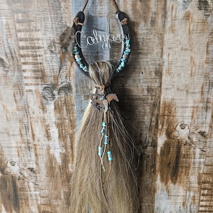 May include: A horseshoe-shaped wall hanging with the name "Calypso" in wire script. The black horseshoe is adorned with turquoise-colored stones and a horsehair tassel. A small metal horse charm hangs below.