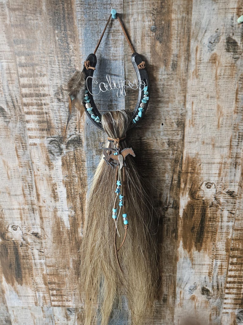 May include: A horseshoe-shaped wall hanging with the name "Calypso" in wire script. The black horseshoe is adorned with turquoise-colored stones and a horsehair tassel. A small metal horse charm hangs below.