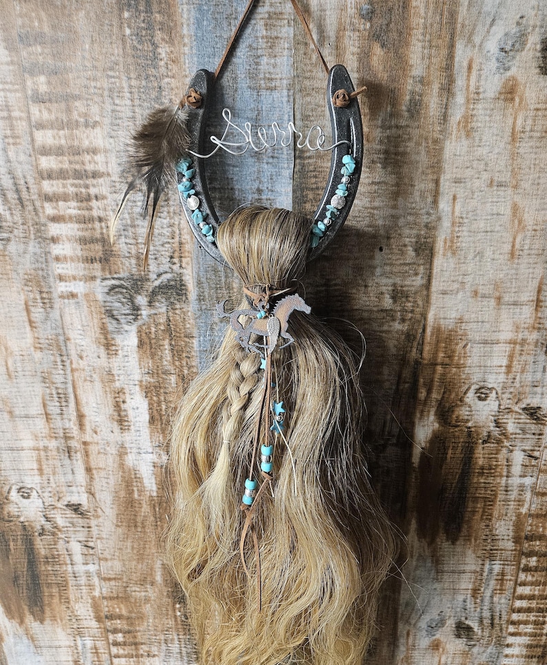 May include: A horseshoe-shaped wall hanging with the name "Sierra" in wire. The horseshoe is decorated with turquoise stones and a feather. A braid of light brown hair is attached, adorned with a horse charm and turquoise beads. The background is a wooden surface.