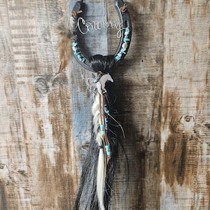 May include: A decorative horseshoe with the word "Cowboy" in cursive script. The horseshoe is adorned with turquoise stones, feathers, and a horse silhouette. Strands of black and white hair hang below, creating a rustic, Western-themed wall hanging.
