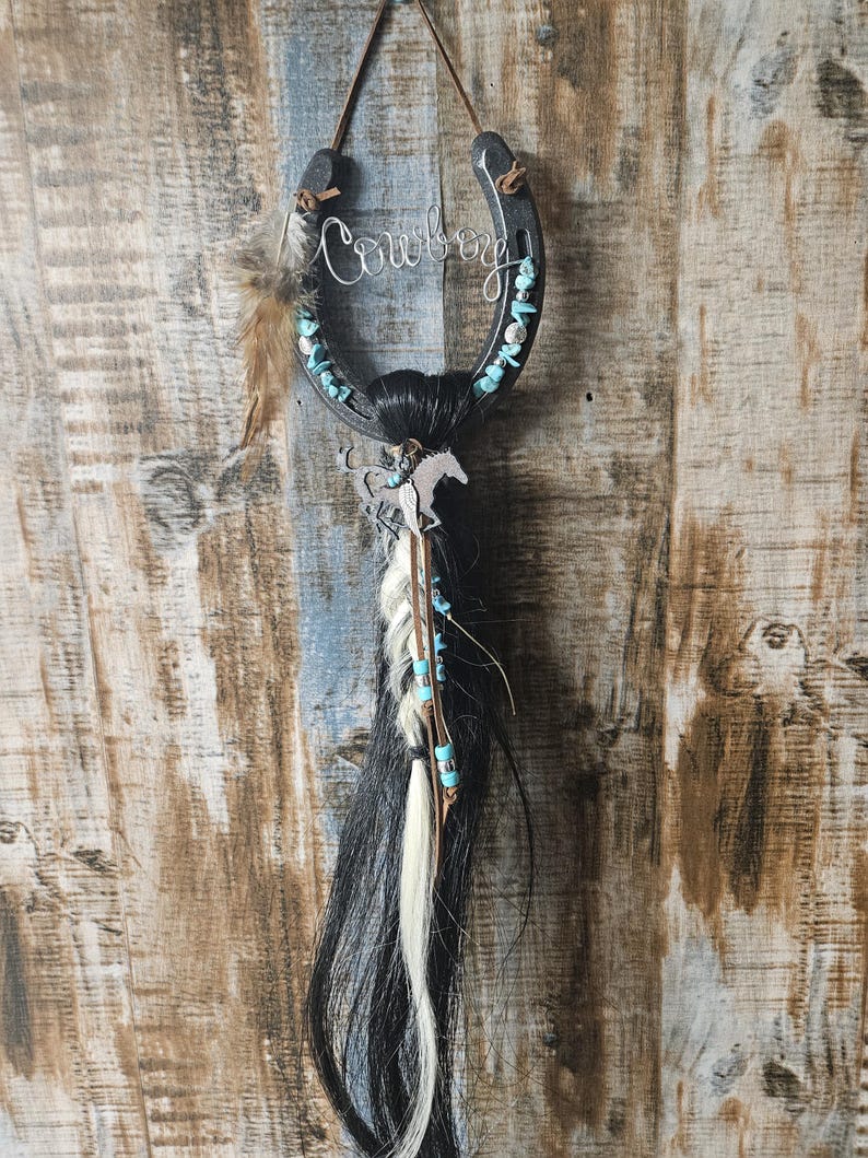 May include: A decorative horseshoe with the word "Cowboy" in cursive script. The horseshoe is adorned with turquoise stones, feathers, and a horse silhouette. Strands of black and white hair hang below, creating a rustic, Western-themed wall hanging.