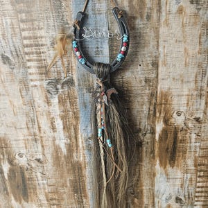 May include: A rustic horseshoe wall hanging with the word "Savior" in wire script. The horseshoe is adorned with red and turquoise beads. Brown horse hair and feathers hang below, with a small metal horse charm.