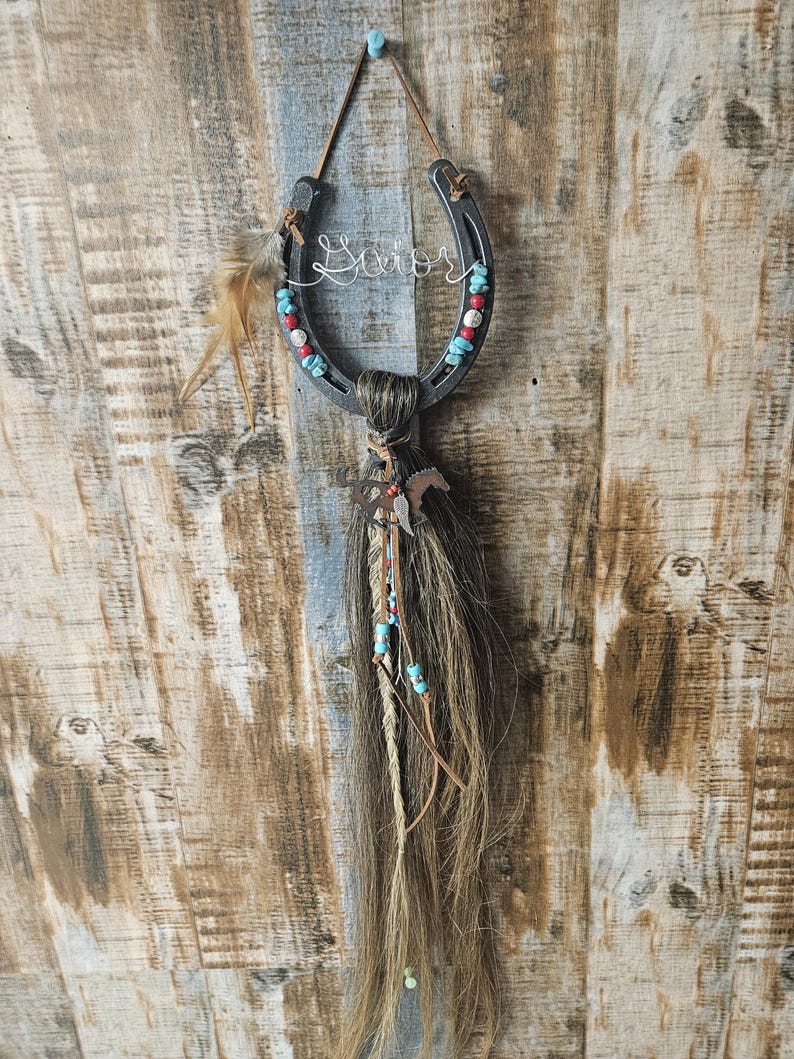 May include: A rustic horseshoe wall hanging with the word "Savior" in wire script. The horseshoe is adorned with red and turquoise beads. Brown horse hair and feathers hang below, with a small metal horse charm.