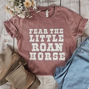 May include: A mauve t-shirt with the text "Fear the Little Roan Horse" printed in white on the front. The shirt is laid out on a wooden surface with a pair of brown boots and blue jeans.