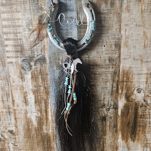 May include: A silver horseshoe with the name "Coal" in wire script, adorned with turquoise beads and a black horsehair tassel. A small horse charm and feathers add to the rustic, Western-themed decor.