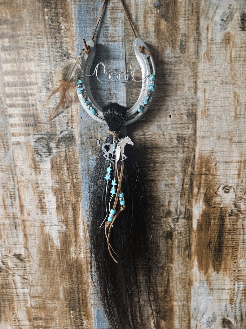 May include: A silver horseshoe with the name "Coal" in wire script, adorned with turquoise beads and a black horsehair tassel. A small horse charm and feathers add to the rustic, Western-themed decor.