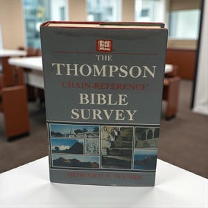 Könnte beinhalten: Ein Hardcover-Buch mit dem Titel "The Thompson Chain-Reference Bible Survey" mit grauem Einband und roten Akzenten. Das Buch enthält den Namen des Autors, Howard A. Hanke, und mehrere kleine Bilder. Das Buch liegt auf einer weißen Oberfläche.
