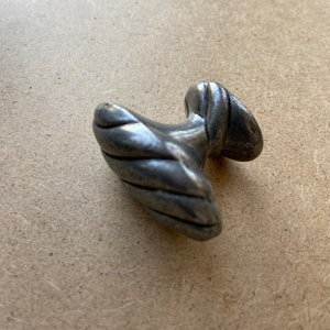 May include: A small, antique-style metal drawer pull with a textured, twisted design. The pull has a rounded handle and a circular base, showing signs of wear and age. The color is a muted silver.