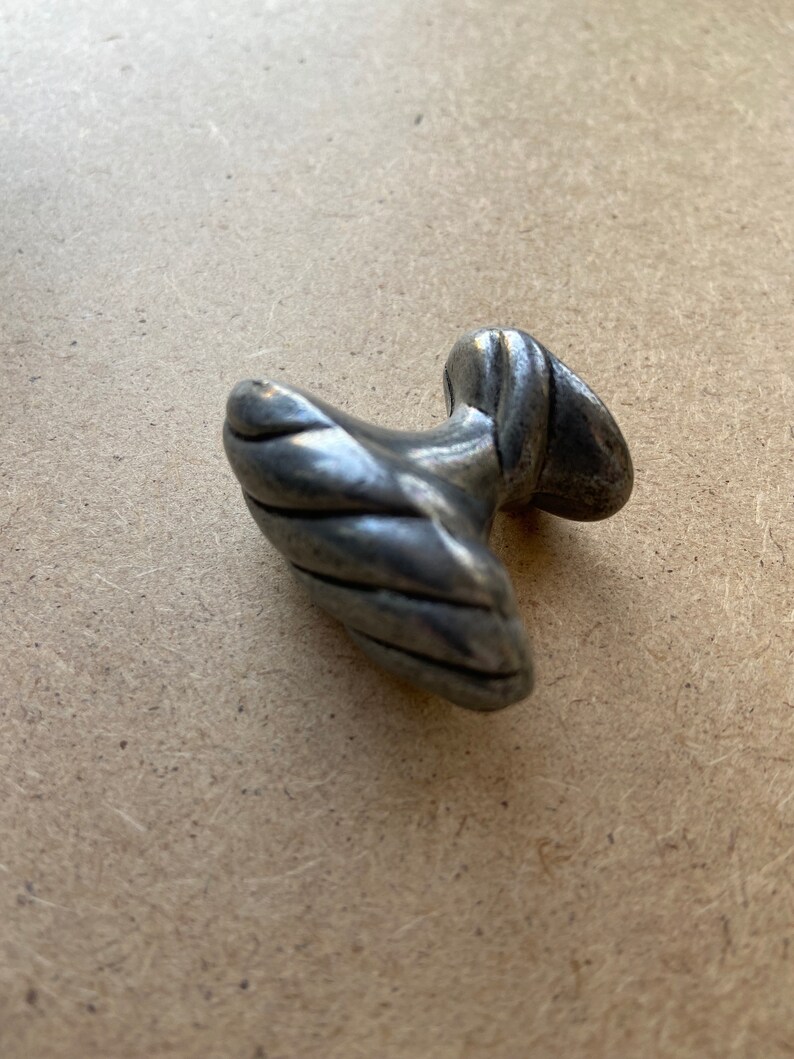 May include: A small, antique-style metal drawer pull with a textured, twisted design. The pull has a rounded handle and a circular base, showing signs of wear and age. The color is a muted silver.