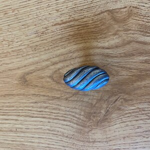 May include: Oval-shaped, antique-style cabinet knob with a textured, ridged surface. The knob has a metallic appearance with a blue-gray hue and is set against a light brown wooden surface.