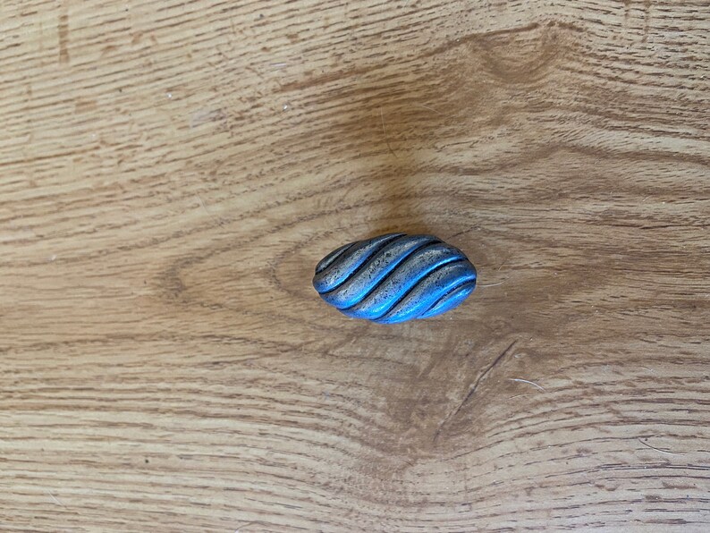 May include: Oval-shaped, antique-style cabinet knob with a textured, ridged surface. The knob has a metallic appearance with a blue-gray hue and is set against a light brown wooden surface.