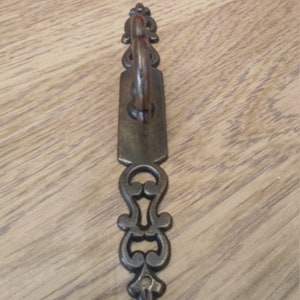 May include: An antique bronze-toned cabinet pull with an ornate design. The pull features a rectangular backplate with a curved handle and decorative flourishes. The hardware is likely for furniture restoration or adding vintage charm.