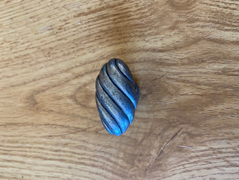 May include: A silver-toned, oval-shaped cabinet knob with a textured, twisted rope design. The hardware is made of metal and is set against a light brown wooden surface.